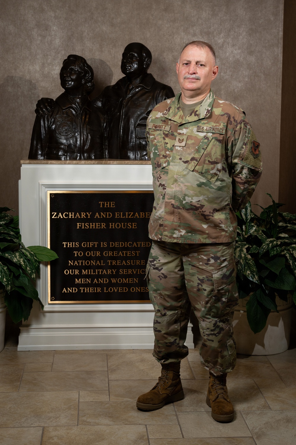 A U.S. Air Force service member in OCP (Operational Camouflage Pattern) uniform stands in front of a dedication plaque and bronze sculpture at the Zachary and Elizabeth Fisher House. The plaque reads 'This gift is dedicated to our greatest national treasure, our military servicemen and women and their loved ones.' The subject appears to be a Technical Sergeant based on visible rank insignia.