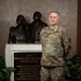 A U.S. Air Force service member in OCP (Operational Camouflage Pattern) uniform stands in front of a dedication plaque and bronze sculpture at the Zachary and Elizabeth Fisher House. The plaque reads 'This gift is dedicated to our greatest national treasure, our military servicemen and women and their loved ones.' The subject appears to be a Technical Sergeant based on visible rank insignia.