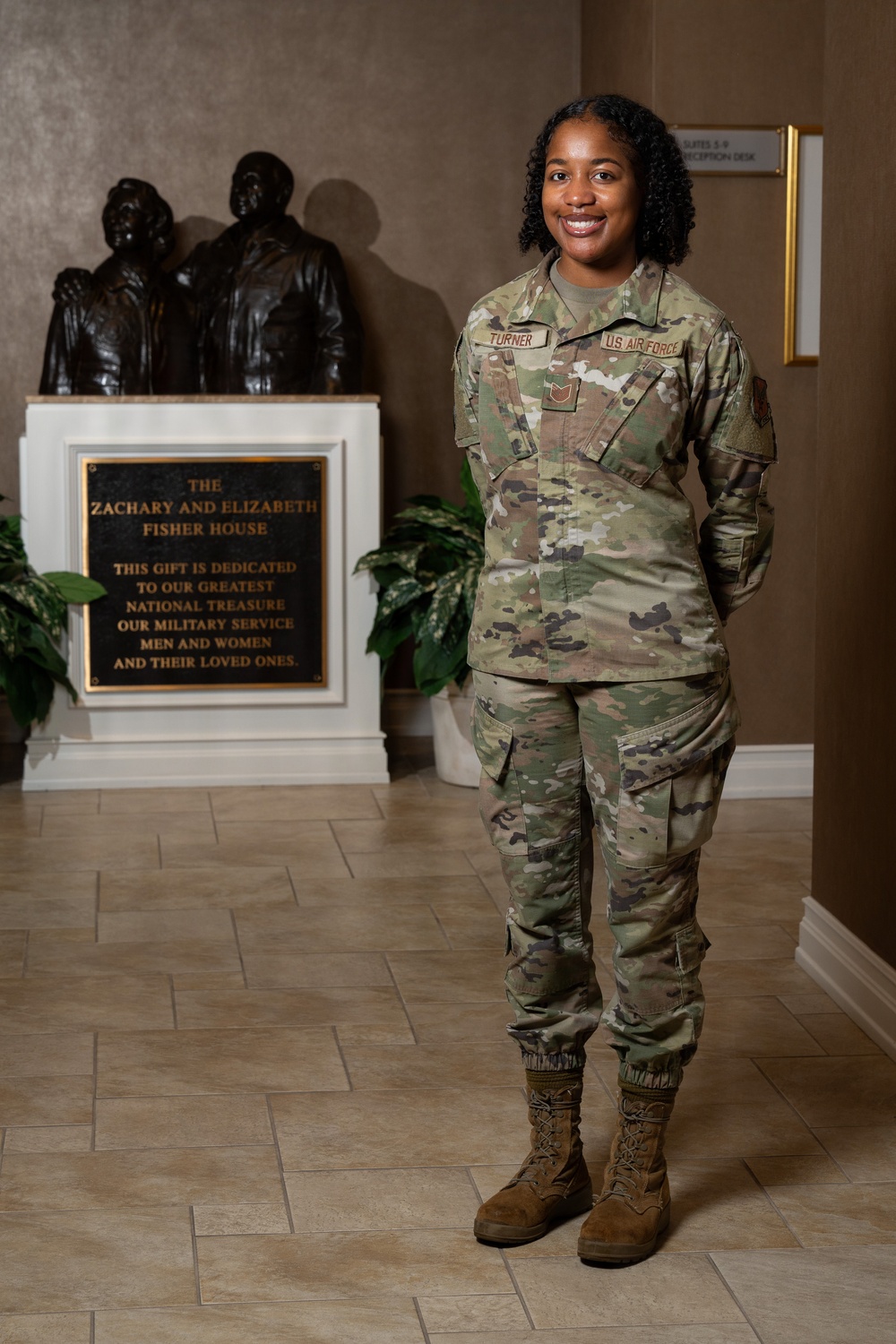 A U.S. Air Force service member named Turner, wearing OCP (Operational Camouflage Pattern) uniform, stands smiling in the lobby of a Fisher House facility. She is posed in front of a bronze dedication plaque and sculpture honoring military service members and their families. The setting is a Fisher House, a nonprofit organization that provides housing for military families during medical treatment.