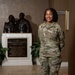 A U.S. Air Force service member named Turner, wearing OCP (Operational Camouflage Pattern) uniform, stands smiling in the lobby of a Fisher House facility. She is posed in front of a bronze dedication plaque and sculpture honoring military service members and their families. The setting is a Fisher House, a nonprofit organization that provides housing for military families during medical treatment.