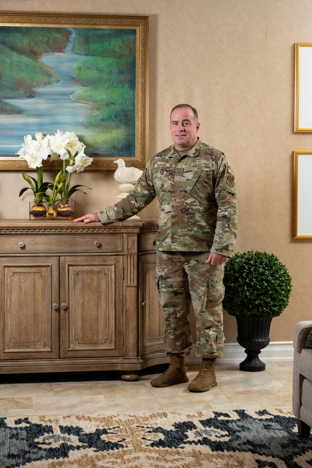A U.S. Air Force service member in OCP (Operational Camouflage Pattern) uniform stands in what appears to be an upscale interior room, posing beside a wooden sideboard. The name tape reads 'BARNAS' and the branch tape reads 'U.S. AIR FORCE.' The setting appears to be a formal portrait or official photograph taken in a well-decorated residential or official quarters environment.