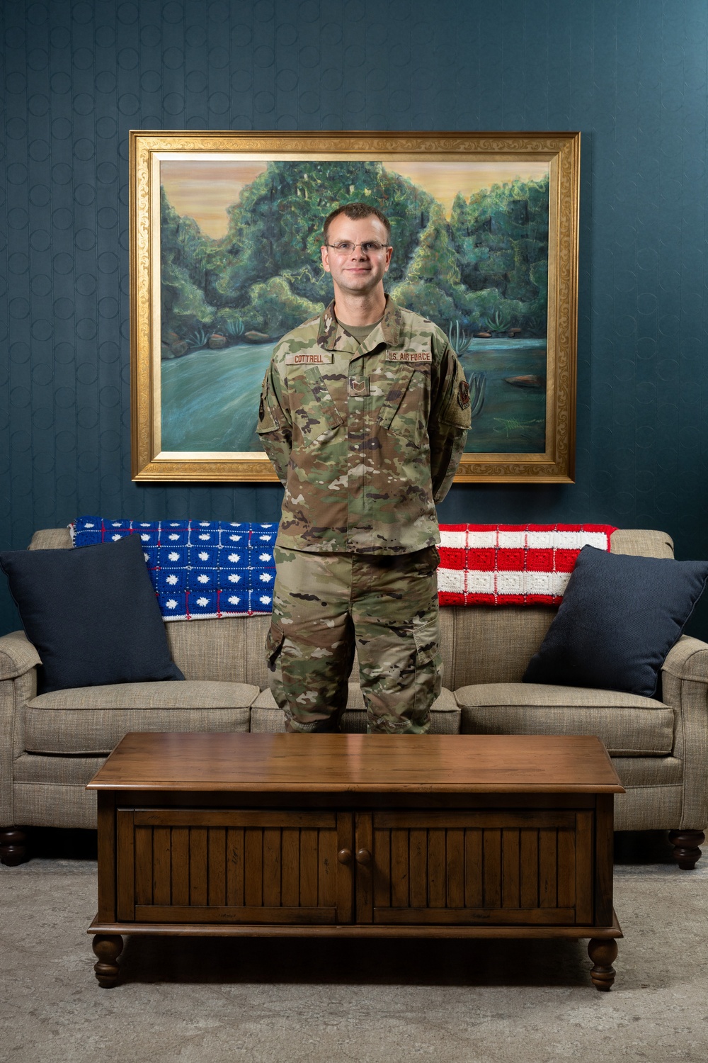 A U.S. Air Force service member named Cottrell stands in a formal portrait setting in front of a large landscape painting with a gold frame. He is wearing the Operational Camouflage Pattern (OCP) uniform and stands behind a wooden coffee table with a sofa visible behind him. The sofa is decorated with crocheted blankets in red, white, and blue American flag patterns.