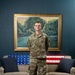 A U.S. Air Force service member named Cottrell stands in a formal portrait setting in front of a large landscape painting with a gold frame. He is wearing the Operational Camouflage Pattern (OCP) uniform and stands behind a wooden coffee table with a sofa visible behind him. The sofa is decorated with crocheted blankets in red, white, and blue American flag patterns.