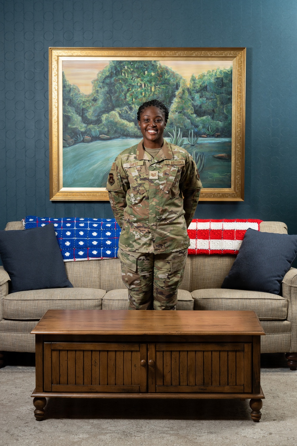 A U.S. Air Force service member in OCP (Operational Camouflage Pattern) uniform stands smiling in what appears to be a formal lounge or common room. The name tape reads 'GUEST' and the branch tape reads 'U.S. AIR FORCE.' The setting features patriotic-themed crocheted blankets draped over a beige sofa, a landscape painting, and teal-colored walls.