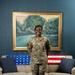 A U.S. Air Force service member in OCP (Operational Camouflage Pattern) uniform stands smiling in what appears to be a formal lounge or common room. The name tape reads 'GUEST' and the branch tape reads 'U.S. AIR FORCE.' The setting features patriotic-themed crocheted blankets draped over a beige sofa, a landscape painting, and teal-colored walls.