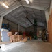 The image shows the interior of a large warehouse or hangar-style building under active construction or renovation. Multiple workers in military-style camouflage uniforms are performing various tasks including working on elevated platforms and installing wall materials. The space contains numerous construction materials including drywall sheets, insulation rolls, and building supplies.