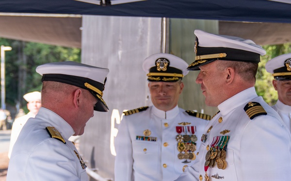 A formal U.S. Navy military ceremony is taking place outdoors, likely a change of command or award presentation. Two senior naval officers in white dress uniforms are shaking hands in the foreground, while two additional officers stand at attention in the background. The setting appears to be a naval installation surrounded by trees.