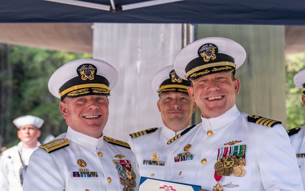 Two senior U.S. Navy officers in white dress uniforms are shaking hands while jointly holding a Legion of Merit certificate during a formal military ceremony. The recipient on the right displays numerous medals and decorations on his uniform. Two additional naval officers stand in the background, and the event appears to be held outdoors under a canopy.