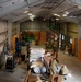A large warehouse or hangar-style building is undergoing active interior renovation or construction. Multiple personnel wearing military-style camouflage uniforms and hard hats are working throughout the space. The scene shows significant construction activity with materials, equipment, and lift platforms in use.