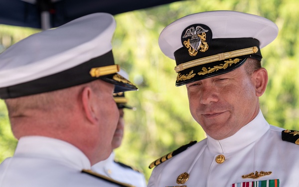 A formal U.S. Navy military ceremony is taking place outdoors, likely a change of command or awards ceremony. Three naval officers in white dress uniforms are visible, with the central figure displaying an extensive array of medals, ribbons, and decorations. The officer facing the camera appears to be a Captain (O-6) based on the four gold stripes on his shoulder boards and the heavily decorated cap with gold oak leaf scrambled eggs.