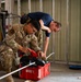 A U.S. Air Force service member in OCP camouflage uniform, identified by name tape as 'ZUNIGA ACUNA,' is kneeling and operating a Milwaukee power tool placed on a red Milwaukee tool case. A second individual in a blue t-shirt is leaning over to observe or assist with the task. The setting appears to be an industrial or maintenance facility with metal wall paneling and various equipment visible in the background.