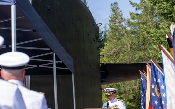 A U.S. Navy officer in white dress uniform delivers a speech at a formal military ceremony from a wooden podium bearing an official seal. The event appears to be an outdoor change of command or commissioning ceremony, with rows of seated Navy personnel in white uniforms and a display of multiple state and military flags. The setting includes a canopy tent, green military equipment in the background, and trees suggesting a naval installation.