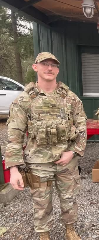A male service member in full MultiCam/OCP camouflage uniform is standing outdoors on a gravel surface. He is wearing a plate carrier/tactical vest with MOLLE pouches and what appears to be a name tape and branch tape visible on the vest. The setting appears to be a training or staging area near a green metal building.