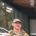 A male service member in full MultiCam/OCP camouflage uniform is standing outdoors on a gravel surface. He is wearing a plate carrier/tactical vest with MOLLE pouches and what appears to be a name tape and branch tape visible on the vest. The setting appears to be a training or staging area near a green metal building.