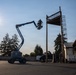A blue articulating boom lift (cherry picker) is elevated with a worker in the basket, positioned near a tall wooden pole and a metal cage ladder structure. The scene appears to be an industrial or training facility yard, photographed against a bright sun creating strong silhouette effects. A second worker is visible on the ground near the base of the metal tower structure.