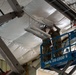 Two individuals in military camouflage uniforms are working at height on a scissor lift, installing or adjusting overhead equipment beneath a white plastic/tarp ceiling liner inside a large metal building. They are using a Genie GS-1930 scissor lift to reach the ceiling area where a suspended light fixture or HVAC unit is being worked on. The setting appears to be a military facility or hangar undergoing construction or renovation.
