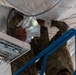 Two military personnel in camouflage uniforms are working overhead on what appears to be a large white tarpaulin or tent structure. They are standing on or near a blue scissor lift or elevated platform, manipulating flexible ducting and fabric material above their heads. One individual is using a flashlight or small light source to illuminate the work area.