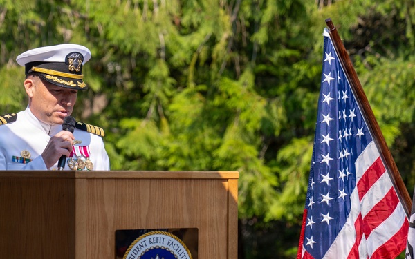 A U.S. Navy officer in dress white uniform is speaking at a podium during an official ceremony at Trident Refit Facility in Bangor, Washington. The officer holds a microphone and wears multiple medals and gold shoulder boards indicating senior rank. The American flag and POW/MIA flag are displayed prominently beside the podium.