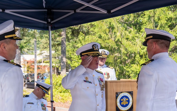 A formal U.S. Navy military ceremony is taking place outdoors under a dark blue canopy tent. A senior naval officer in white dress uniform is rendering a salute at a wooden podium bearing an official Navy seal/emblem. Several other naval officers in white dress uniforms are present, with flags including a POW/MIA flag visible in the background.