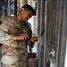 A U.S. Air Force service member named Zuniga Acuna, wearing OCP (Operational Camouflage Pattern) uniform and safety glasses, is performing construction or renovation work on metal wall framing. The airman appears to be fastening or securing metal studs/framing components in what looks like an active construction site. The setting suggests a humanitarian assistance, civil engineering, or base construction project.