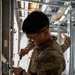 A U.S. Air Force service member wearing OCP (Operational Camouflage Pattern) uniform with an 'HVACR' specialty patch is performing maintenance or installation work using a screwdriver on what appears to be an electrical or mechanical component mounted on a wall frame. The setting is an active construction or renovation site with exposed metal wall studs and ceiling framing visible. A second service member in the same uniform can be partially seen working in the background.