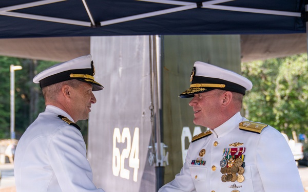 A formal U.S. Navy military ceremony is taking place outdoors under a dark canopy. Two naval officers in white dress uniforms are shaking hands, with a third officer visible in the background to the right. The number '624' is visible on a metallic structure behind them, suggesting this may be a change of command or commissioning ceremony.