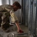 A military service member in MultiCam/OCP camouflage uniform is performing construction or renovation work, applying mortar or adhesive to a floor surface using a notched trowel. The individual is kneeling on the floor near a metal door frame or structural framing, wearing protective safety glasses. The setting appears to be an interior construction or building improvement project.