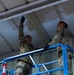 Two military personnel in camouflage uniforms and white hard hats are working on an elevated scissor lift platform, measuring the ceiling or upper wall area of a metal building. One individual is reaching upward holding a yellow tape measure against the ceiling while the other assists. They appear to be conducting construction, inspection, or renovation work inside a metal-paneled structure.