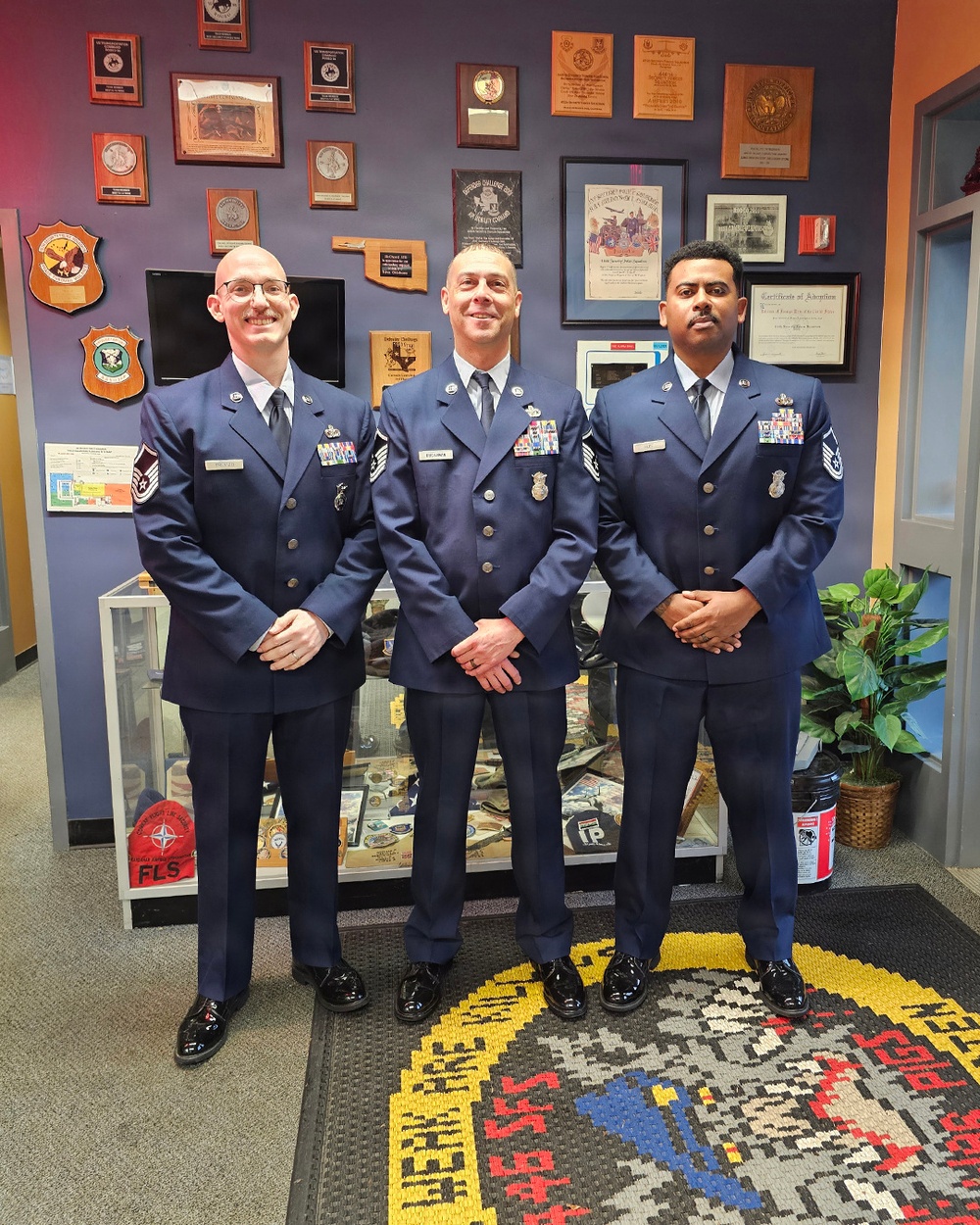Three U.S. Air Force personnel in dress blue uniforms are posing together in what appears to be a military unit office or hallway. The wall behind them is decorated with numerous plaques, awards, certificates, and unit insignia. All three individuals are wearing service dress uniforms with ribbons, badges, and rank insignia visible.
