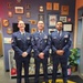 Three U.S. Air Force personnel in dress blue uniforms are posing together in what appears to be a military unit office or hallway. The wall behind them is decorated with numerous plaques, awards, certificates, and unit insignia. All three individuals are wearing service dress uniforms with ribbons, badges, and rank insignia visible.