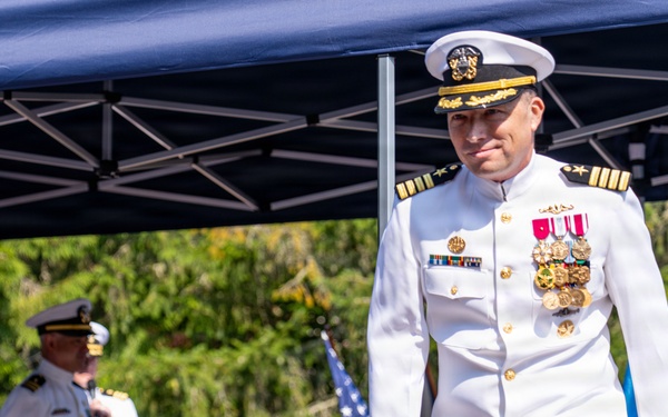 A U.S. Navy officer in white dress uniform adorned with multiple medals and ribbons walks forward at what appears to be a formal military ceremony, likely a change of command or retirement event. In the background, another naval officer stands at a podium beneath a blue canopy tent, with an American flag and what appears to be a unit or command flag visible. The outdoor setting features lush green trees and a wooden railing structure.