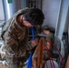 Two military personnel in camouflage uniforms are performing plumbing or mechanical work in what appears to be a utility or mechanical room. The foreground soldier, identified by name tape as 'ROBBINS,' is bent over working with blue flexible tubing near wooden framing. A second service member works in the background near copper and other piping infrastructure.