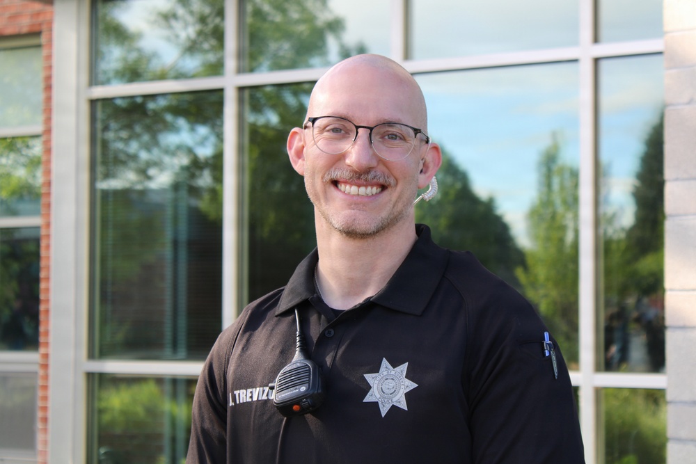 A smiling bald man wearing glasses and a black law enforcement uniform polo shirt is photographed outdoors in front of a large glass window building. His name tag reads 'J. TREVIZ...' and he has a police/sheriff badge on his sleeve along with a radio microphone clipped to his shirt. The image appears to be a professional or semi-professional portrait photo taken in good lighting conditions.