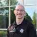 A smiling bald man wearing glasses and a black law enforcement uniform polo shirt is photographed outdoors in front of a large glass window building. His name tag reads 'J. TREVIZ...' and he has a police/sheriff badge on his sleeve along with a radio microphone clipped to his shirt. The image appears to be a professional or semi-professional portrait photo taken in good lighting conditions.