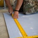 A person wearing military camouflage (OCP/MultiCam pattern) uniform is kneeling on the floor, measuring or marking a sheet of drywall or similar building material. They are using a yellow carpenter's square or ruler to take measurements on the flat panel. The person is wearing a ring on their right hand and a black bracelet on their wrist.