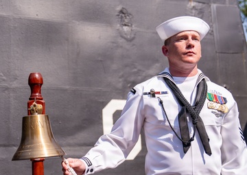 A U.S. Navy sailor in dress white uniform stands beside a brass ship's bell mounted on a wooden stand, appearing to ring or hold the bell's lanyard during a formal ceremony. The sailor displays multiple service medals and ribbons on his chest, along with a black neckerchief, indicating a formal naval occasion. The background shows the gray hull of a naval vessel, suggesting this ceremony is taking place dockside.