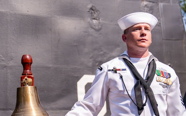A U.S. Navy sailor in dress white uniform stands beside a brass ship's bell mounted on a wooden stand, appearing to ring or hold the bell's lanyard during a formal ceremony. The sailor displays multiple service medals and ribbons on his chest, along with a black neckerchief, indicating a formal naval occasion. The background shows the gray hull of a naval vessel, suggesting this ceremony is taking place dockside.