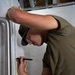 A young man wearing military-style camouflage clothing and safety glasses is marking measurements on a white surface using a tape measure and a marker. He is leaning forward intently, using one hand to hold the marker at a specific measurement point while his other arm braces against the surface. The setting appears to be an active construction or renovation environment.