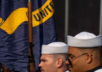 The image shows United States Navy personnel in a formal color guard or ceremonial formation. Sailors are dressed in dress blue and white uniforms, carrying rifles and a Navy flag with visible text 'UNITE' (United States Navy). The scene appears to be an official military ceremony or parade.