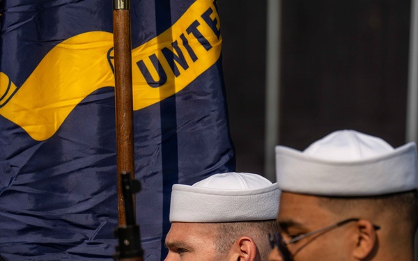 The image shows United States Navy personnel in a formal color guard or ceremonial formation. Sailors are dressed in dress blue and white uniforms, carrying rifles and a Navy flag with visible text 'UNITE' (United States Navy). The scene appears to be an official military ceremony or parade.