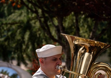 A U.S. Navy sailor in dress white uniform is playing a tuba during an outdoor band performance. The sailor wears a white dixie cup cover and has service ribbons visible on the chest. Music stands with sheet music are positioned in front of the performers.
