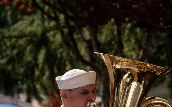 A U.S. Navy sailor in dress white uniform is playing a tuba during an outdoor band performance. The sailor wears a white dixie cup cover and has service ribbons visible on the chest. Music stands with sheet music are positioned in front of the performers.