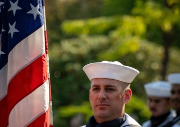 A U.S. Navy sailor in dress white uniform stands at attention holding an American flag with gold fringe, serving as part of a color guard. The sailor displays a petty officer first class rating badge on the right sleeve along with a blue aiguillette, indicating ceremonial duty. At least one additional sailor in matching dress whites is visible in the background.