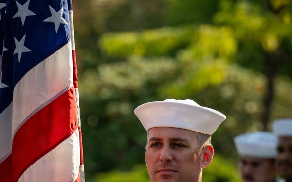 A U.S. Navy sailor in dress white uniform stands at attention holding an American flag with gold fringe, serving as part of a color guard. The sailor displays a petty officer first class rating badge on the right sleeve along with a blue aiguillette, indicating ceremonial duty. At least one additional sailor in matching dress whites is visible in the background.