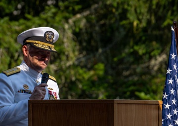 A senior U.S. Navy officer in white dress uniform is speaking at a podium during an official ceremony at Trident Refit Facility, Bangor, Washington. The officer is holding a microphone and appears to be delivering a speech or remarks at what is likely a formal military event. An American flag is displayed to the right of the podium.