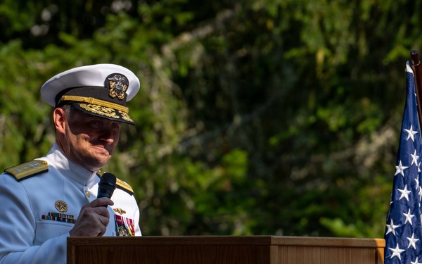 A senior U.S. Navy officer in white dress uniform is speaking at a podium during an official ceremony at Trident Refit Facility, Bangor, Washington. The officer is holding a microphone and appears to be delivering a speech or remarks at what is likely a formal military event. An American flag is displayed to the right of the podium.