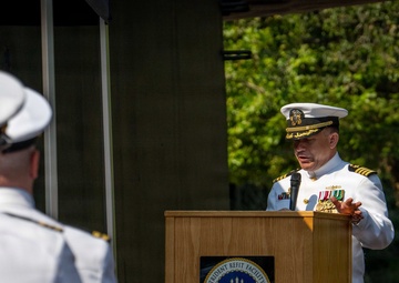 A U.S. Navy officer in dress white uniform is delivering a speech at a podium bearing the seal of Trident Refit Facility, Bangor, Washington. A formation of Navy personnel in white uniforms stands at attention in the foreground, while military color guard flags are displayed to the right. The setting appears to be an outdoor military ceremony, likely a change of command or official commissioning event.