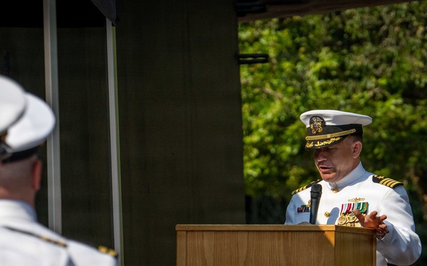 A U.S. Navy officer in dress white uniform is delivering a speech at a podium bearing the seal of Trident Refit Facility, Bangor, Washington. A formation of Navy personnel in white uniforms stands at attention in the foreground, while military color guard flags are displayed to the right. The setting appears to be an outdoor military ceremony, likely a change of command or official commissioning event.