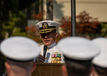 A senior U.S. Navy officer in dress white uniform is delivering a speech at a podium during a formal military ceremony. The podium bears a seal reading 'Trident Refit Facility,' indicating this is an official Navy installation event. An audience of uniformed sailors in white covers (hats) is seated or standing with their backs to the camera.