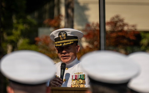 A senior U.S. Navy officer in dress white uniform is delivering a speech at a podium during a formal military ceremony. The podium bears a seal reading 'Trident Refit Facility,' indicating this is an official Navy installation event. An audience of uniformed sailors in white covers (hats) is seated or standing with their backs to the camera.